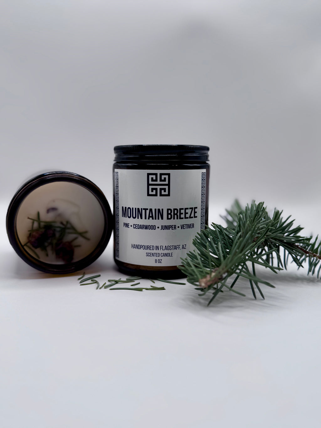 Candle jar labeled 'Mountain Breeze' with a decorative label, surrounded by greenery on a light background.