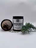 Candle jar labeled 'Mountain Breeze' with a decorative label, surrounded by greenery on a light background.